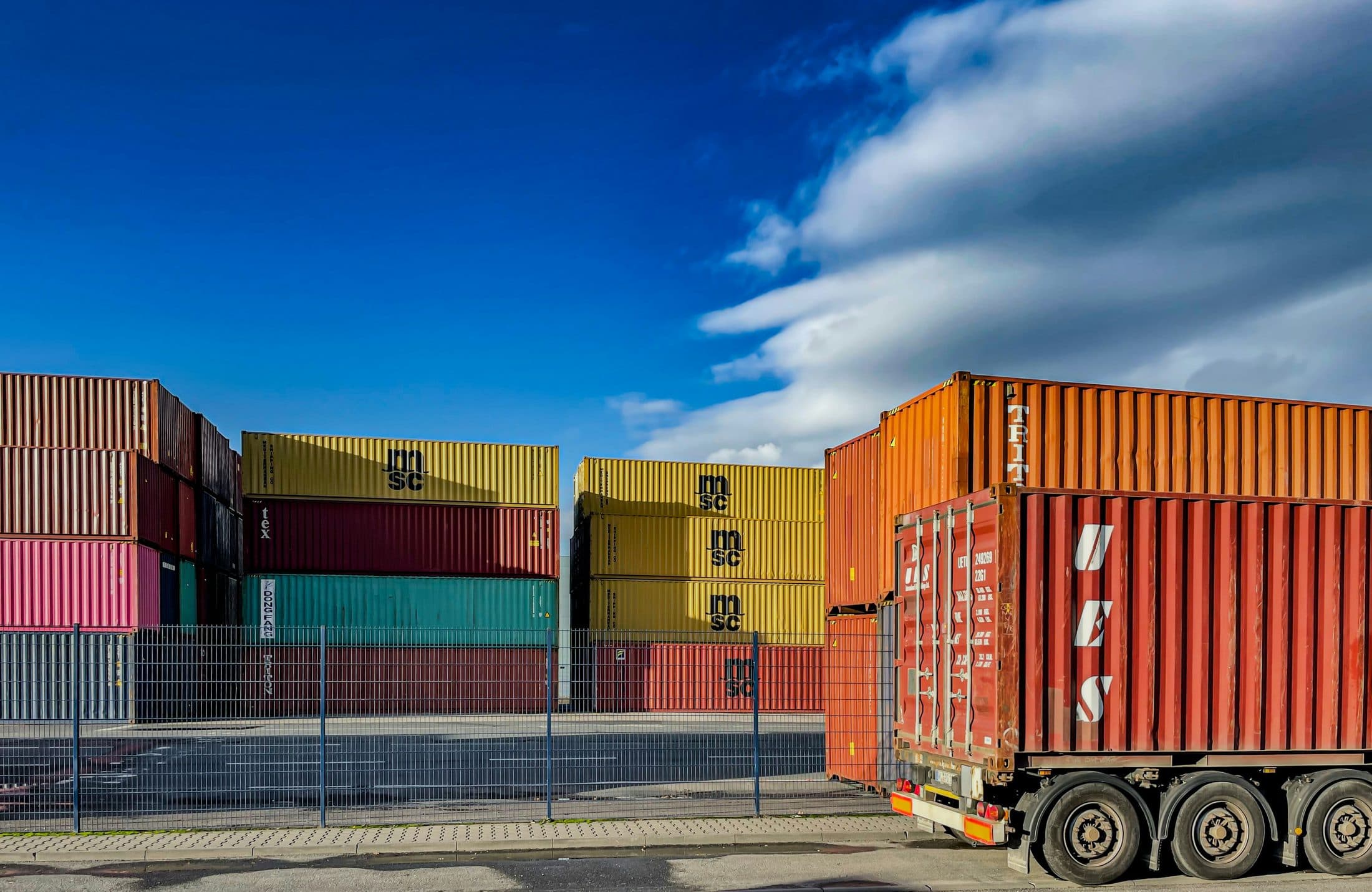A truck loaded with a container beside stacked shipping containers